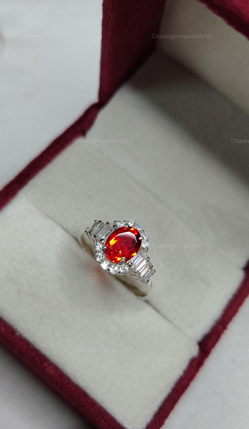 K&ouml;nnte beinhalten: Silberring mit einem gro&szlig;en, oval geschliffenen, feuerroten Edelstein als Mittelpunkt. Der Edelstein ist von einem Kranz aus kleinen, klaren Steinen umgeben. Der Ring hat auch rechteckige, klare Steine auf jeder Seite.