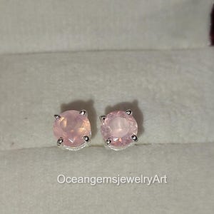 Puede incluir: Un par de pendientes de botón redondos de piedra preciosa rosa claro. Las piedras preciosas están engastadas en metal plateado. Los pendientes se muestran en una caja de joyería gris claro. El texto "OceangemsjewelryArt" es visible.