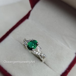 May include: A silver ring with a large, oval-cut emerald gemstone as the centerpiece. The ring is set with smaller, clear gemstones and has a decorative leaf design on the sides. The ring is displayed in a red velvet jewelry box. The text "OceangemsjewelryArt" is visible.