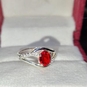 May include: A silver ring with a large, oval, bright red gemstone as the focal point. The band features small, clear accent stones. The ring is displayed in a red and white jewelry box, showcasing the ring's design and color.
