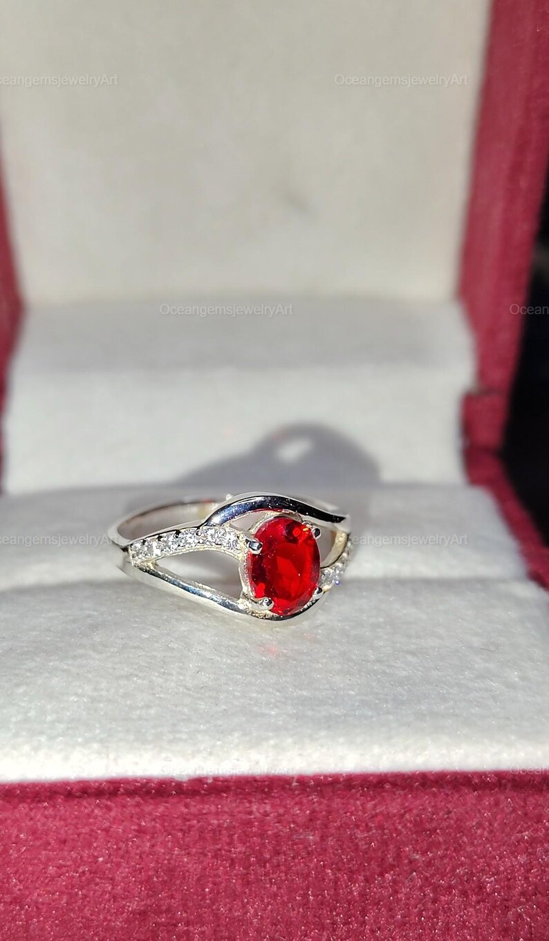 May include: A silver ring with a large, oval, bright red gemstone as the focal point. The band features small, clear accent stones. The ring is displayed in a red and white jewelry box, showcasing the ring's design and color.