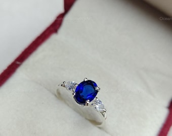 May include: A silver ring with a large, oval, deep blue gemstone as the centerpiece, flanked by two smaller, pear-shaped, clear gemstones. The ring is set inside a red jewelry box.