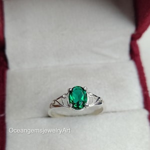 Puede incluir: Anillo de plata con una piedra preciosa ovalada de color verde esmeralda. El anillo tiene una banda decorativa con detalles de filigrana. Se presenta en un joyero de terciopelo rojo. El texto "OceangemsjewelryArt" es visible.