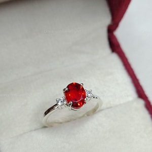 Puede incluir: Un anillo de plata con una gran gema roja ovalada como pieza central. Dos gemas claras más pequeñas flanquean la piedra principal. El anillo se presenta en un joyero forrado de terciopelo.