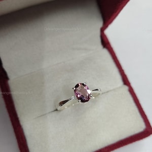 May include: A silver ring featuring a large, oval-cut, pink gemstone. The ring has a simple, elegant design and is presented in a red jewellery box with a white interior. The ring is a piece of jewellery.