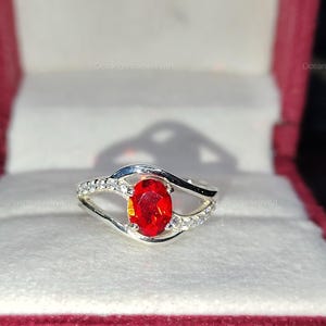 May include: A silver ring with a large, oval-cut, fiery red gemstone as the focal point. The band features a split design with small, clear accent stones. The ring is displayed in a red jewelry box, highlighting its vibrant color and design.