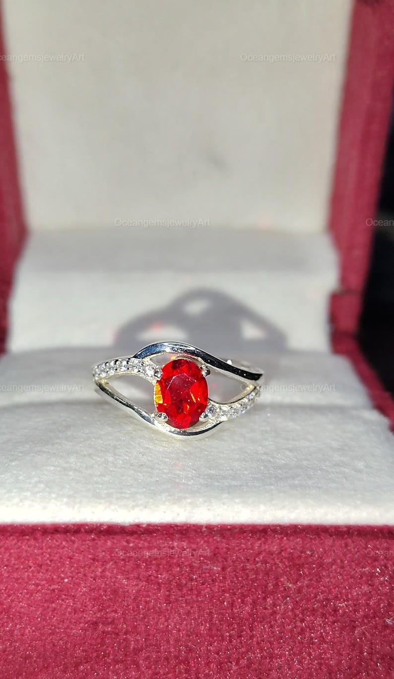 May include: A silver ring with a large, oval-cut, fiery red gemstone as the focal point. The band features a split design with small, clear accent stones. The ring is displayed in a red jewelry box, highlighting its vibrant color and design.