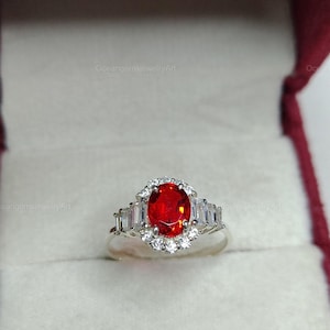 Può includere: Un anello d'argento con una grande pietra rossa ovale al centro, circondata da pietre bianche più piccole. L'anello presenta un design a alone con pietre bianche rettangolari su ogni lato. L'anello è esposto in un portagioie bordeaux.