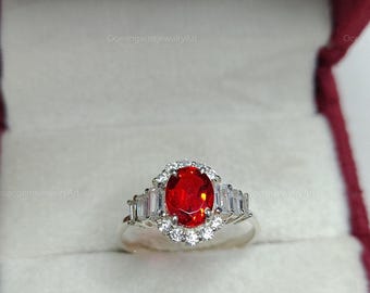 May include: A silver ring featuring a large, oval, red gemstone, encircled by smaller white stones. The ring has a halo design with rectangular white stones on either side. The ring is presented in a maroon jewellery box.