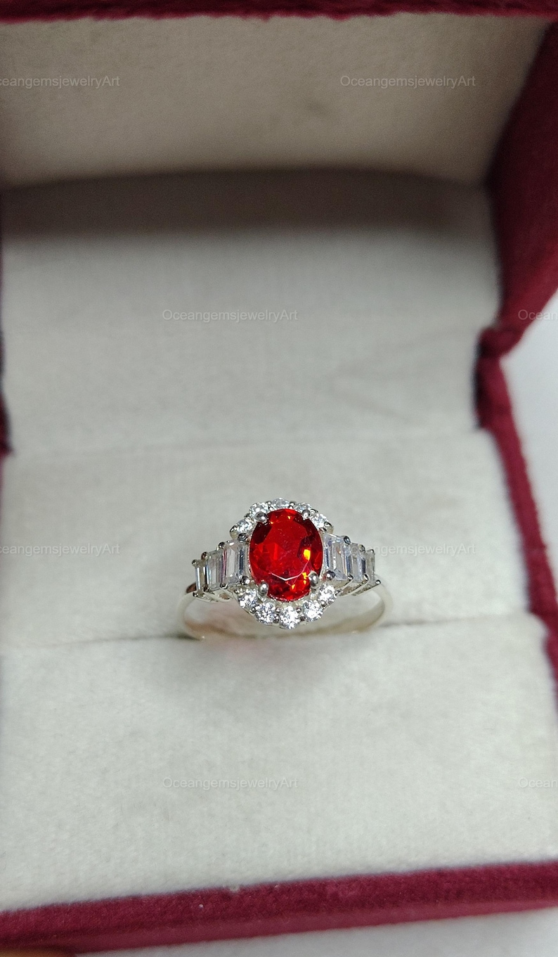 K&ouml;nnte beinhalten: Ein silberner Ring mit einem gro&szlig;en, ovalen, roten Edelstein in der Mitte, umgeben von kleineren wei&szlig;en Steinen. Der Ring hat ein Halo-Design mit zus&auml;tzlichen rechteckigen wei&szlig;en Steinen auf jeder Seite. Der Ring wird in einer weinroten Schmuckschatulle pr&auml;sentiert.