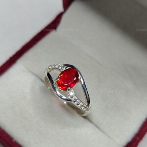 May include: A silver ring with a large, oval-cut, fiery red gemstone as the focal point. The band features a split design with small, clear accent stones. The ring is presented in a burgundy jewelry box.
