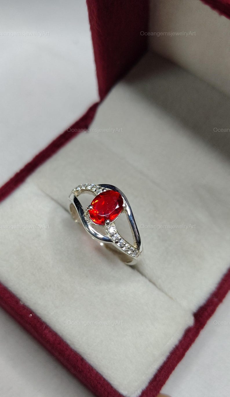 May include: A silver ring with a large, oval-cut, fiery red gemstone as the focal point. The band features a split design with small, clear accent stones. The ring is presented in a burgundy jewelry box.