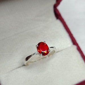 May include: A silver ring with a vibrant red oval gemstone, set in a classic prong setting. The ring features a delicate band and small accent stones on either side of the main stone. The ring is displayed in a red jewelry box.