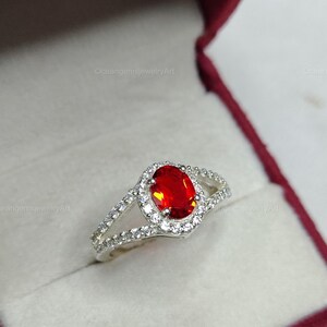 Peut inclure: Bague en argent ornée d'une grande pierre rouge ovale au centre, entourée d'un halo de petites pierres claires. Le corps de la bague se divise en deux, chacune sertie de pierres claires. La bague est présentée dans un écrin rouge.