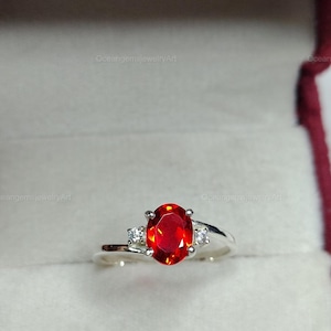 Può includere: Un anello d'argento con una grande gemma rossa ovale come elemento centrale. Due gemme più piccole e chiare affiancano la pietra principale. L'anello è presentato su uno sfondo neutro, evidenziando il design del gioiello.