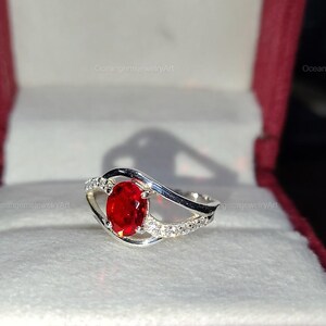 May include: A silver ring with a large oval, bright red gemstone as the focal point. The band features a wave design with small, clear accent stones. The ring is displayed in a red jewelry box.