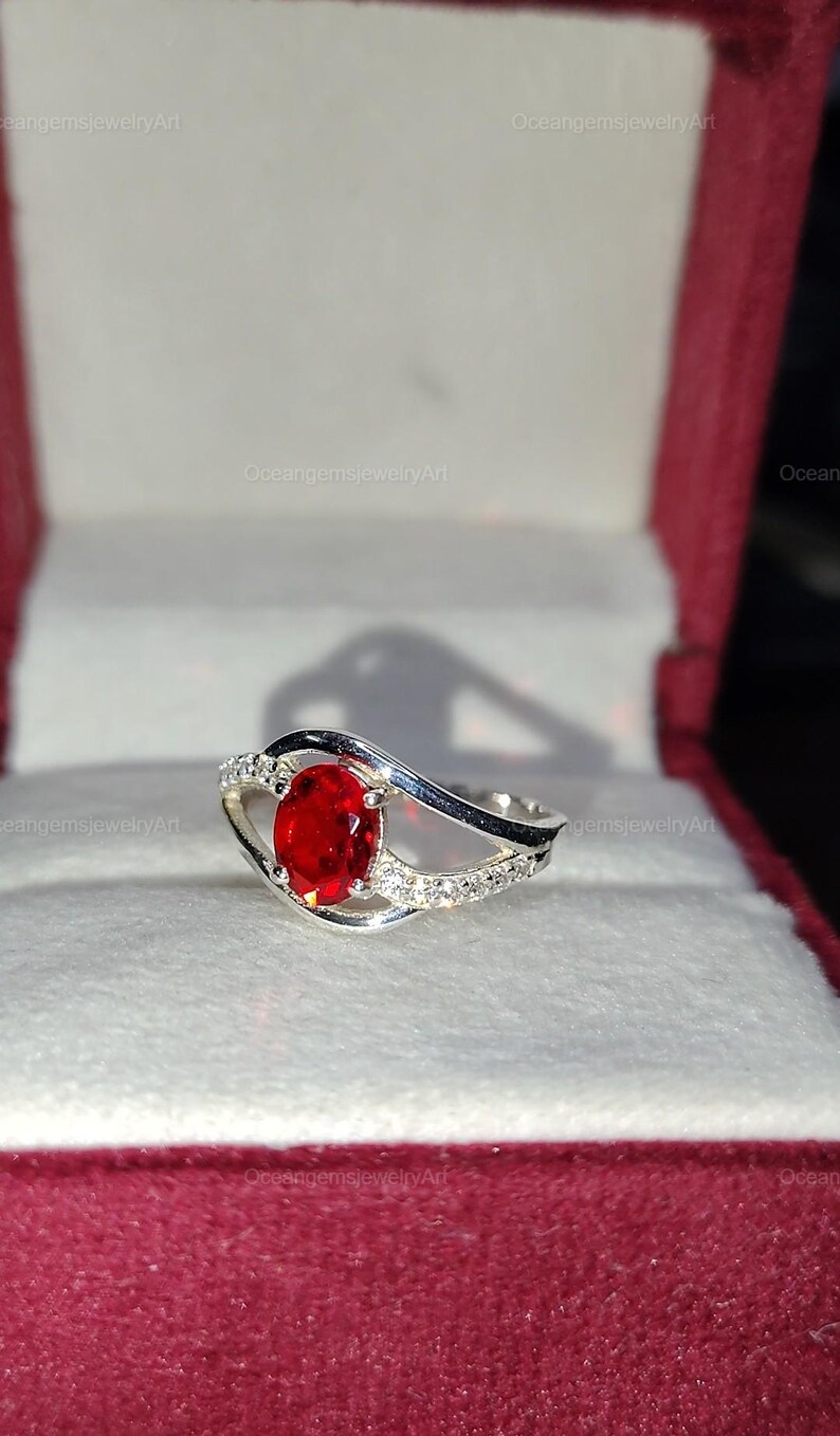 May include: A silver ring with a large oval, bright red gemstone as the focal point. The band features a wave design with small, clear accent stones. The ring is displayed in a red jewelry box.
