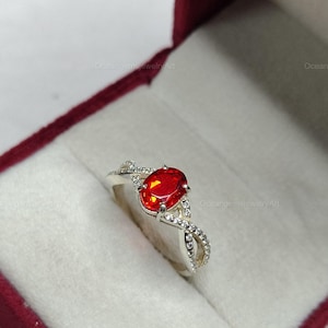 May include: A silver ring with a large, oval-shaped, fiery red gemstone as the centerpiece. The band features a delicate, intertwined design with small, clear accent stones, set against a white background inside a red jewelry box.