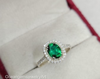 May include: A silver ring featuring an oval emerald gemstone, encircled by a halo of smaller, clear stones. The ring is presented in a red jewellery box. The text "OceangemsjewelryArt" is visible.