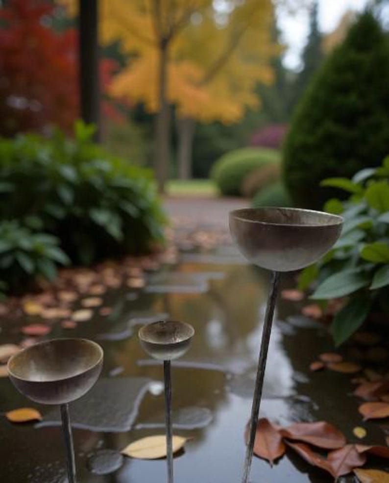 Puede incluir: Tres estacas de jard&iacute;n met&aacute;licas en forma de cuenco de diferentes alturas se mantienen erguidas. Cada estaca cuenta con un tallo delgado y fino que sostiene un cuenco plateado, liso y redondeado, dise&ntilde;ado para recoger agua o peque&ntilde;os objetos.