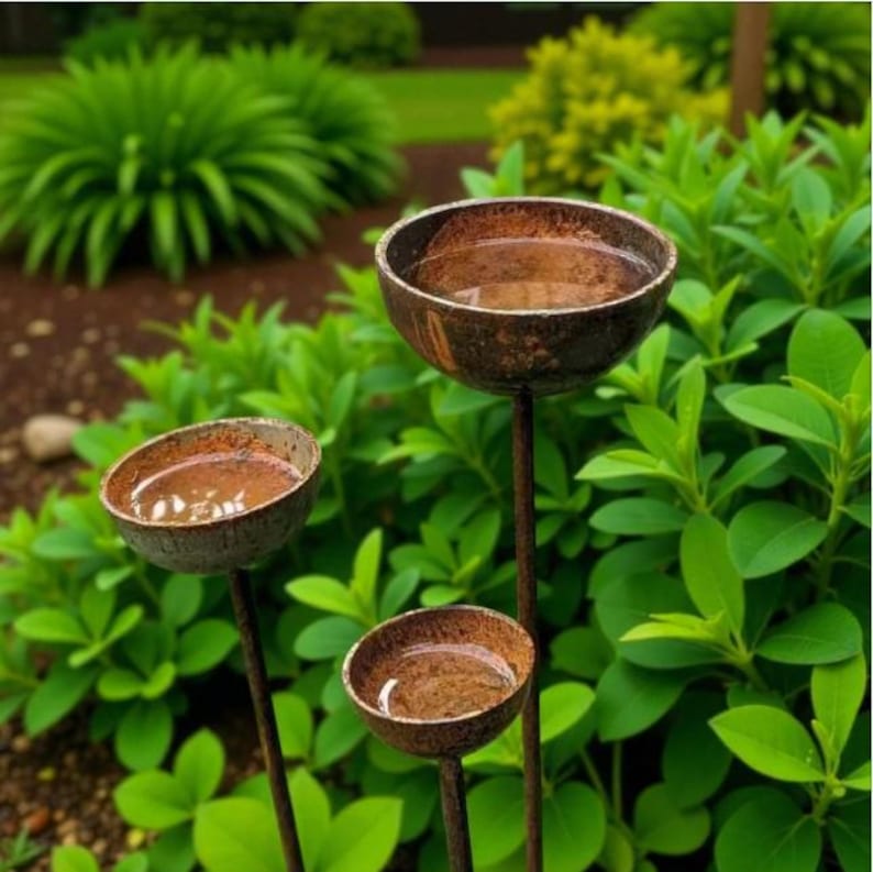 Puede incluir: Tres estacas de jard&iacute;n con forma de cuenco circular y acabado met&aacute;lico en color marr&oacute;n oscuro desgastado. Cada cuenco contiene agua y est&aacute; montado sobre una fina varilla met&aacute;lica vertical, creando una disposici&oacute;n escalonada.