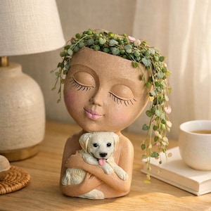 Op de afbeelding: Een beige gekleurde hars plantenbak in de vorm van een menselijk hoofd met gesloten ogen en roze wangen, die een kleine witte puppy in de armen houdt. De bovenkant van het hoofd dient als houder voor een lantaarnplantje met groene en roze blaadjes.