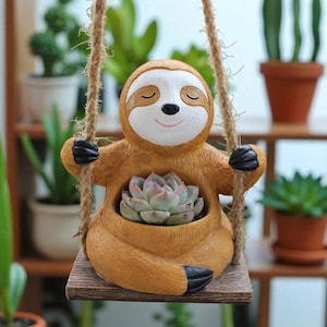 Puede incluir: Una maceta marrón con forma de perezoso con una cara sonriente y ojos cerrados, que sostiene una planta suculenta. La maceta está suspendida de un columpio de madera por una cuerda. El perezoso tiene garras negras y una cara blanca.