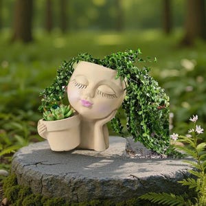 May include: A beige ceramic planter shaped like a woman's head with closed eyes and pink lips. The planter holds a small potted succulent and is adorned with trailing green plants, creating a whimsical, natural look. The planter is resting on a stone.