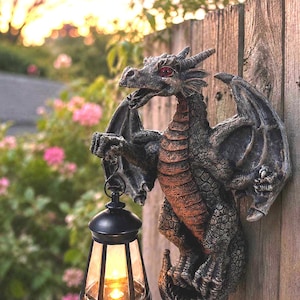 May include: A wall-mounted dragon sculpture featuring a textured gray and copper-toned body with red eyes. The dragon holds a black lantern in one claw, which contains a glowing light source, creating a detailed decorative piece for outdoor use.