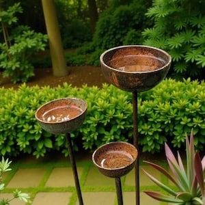 Puede incluir: Tres bebederos para p&aacute;jaros de metal, de forma circular y color marr&oacute;n oscuro, montados sobre finas estacas met&aacute;licas verticales. Cada cuenco est&aacute; lleno de agua, reflejando el entorno. Las estructuras est&aacute;n dispuestas a diferentes alturas.