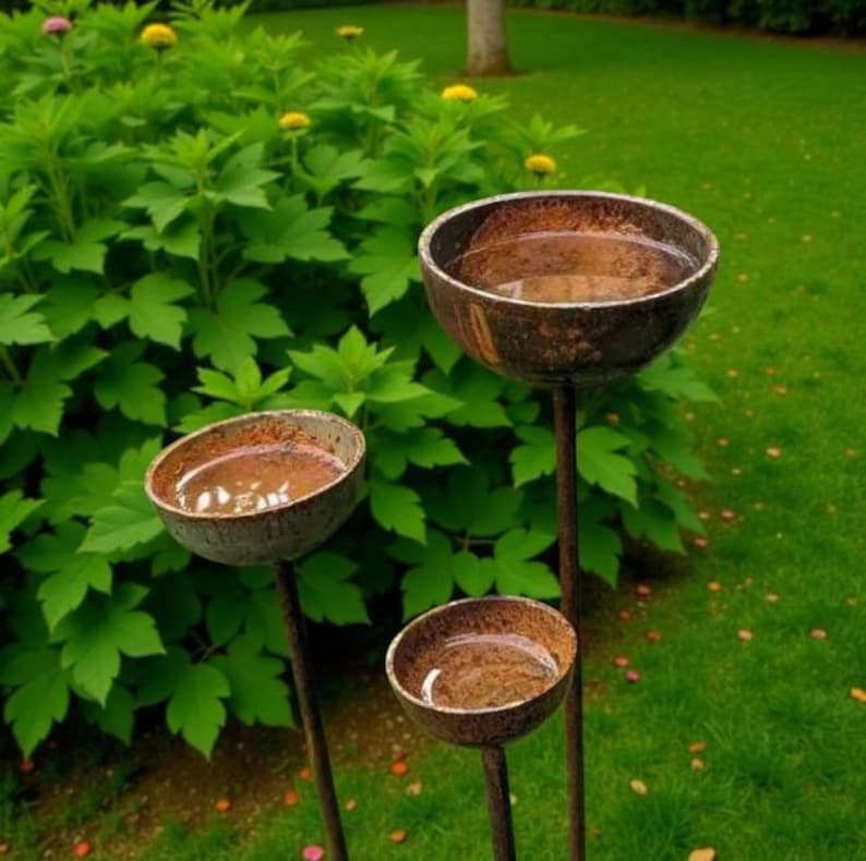 Puede incluir: Tres estacas de jard&iacute;n con forma de cuenco circular, hechas de metal oscuro y desgastado con un acabado oxidado y texturizado. Cada cuenco est&aacute; montado sobre una varilla met&aacute;lica vertical delgada y lleno de agua, reflejando el follaje verde.