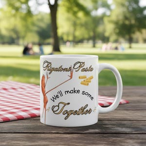 May include: White ceramic mug with the words "Rigatoni Pasta" and "We'll make some Together" in a decorative font. The mug features a cartoon illustration of a rigatoni pasta character. The background is a blurred outdoor scene.