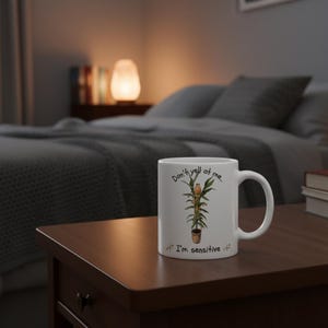 May include: White ceramic mug with a plant illustration and the text "Don't yell at me" and "I'm sensitive". The mug has a curved handle and is sitting on a brown wooden surface.