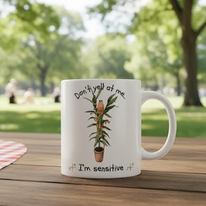 May include: White ceramic mug with a humorous design. The mug features the text "Don't yell at me... I'm sensitive" along with an illustration of a cartoon frog on a plant. The mug has a curved handle and is suitable for hot or cold beverages.