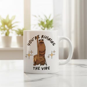 May include: White ceramic mug with a humorous illustration of a brown hand giving the middle finger, wearing sunglasses. The text on the mug reads "YOU'RE RUINING THE VIBE" in black lettering, with gold star accents.