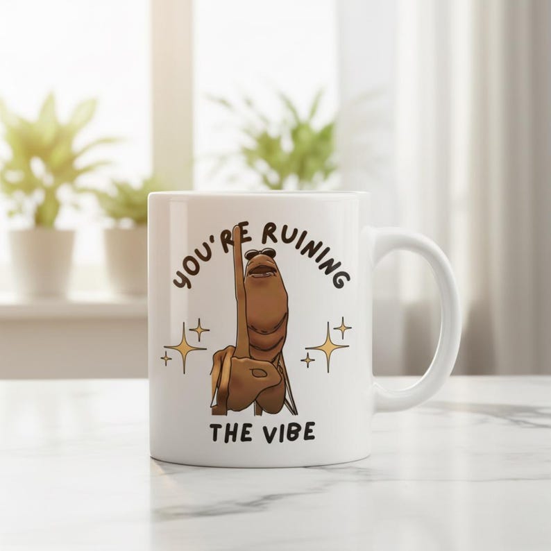 May include: White ceramic mug with a humorous illustration of a brown hand giving the middle finger, wearing sunglasses. The text on the mug reads "YOU'RE RUINING THE VIBE" in black lettering, with gold star accents.
