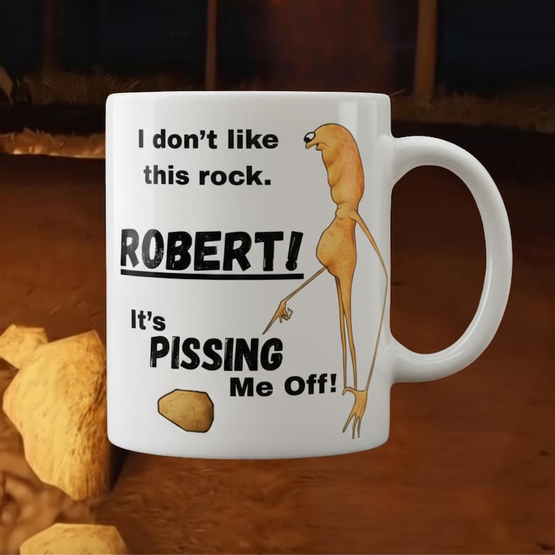 May include: White ceramic mug with a humorous design. The mug features the text "I don't like this rock. ROBERT! It's PISSING Me Off!" along with a cartoon illustration of a creature pointing at a rock.