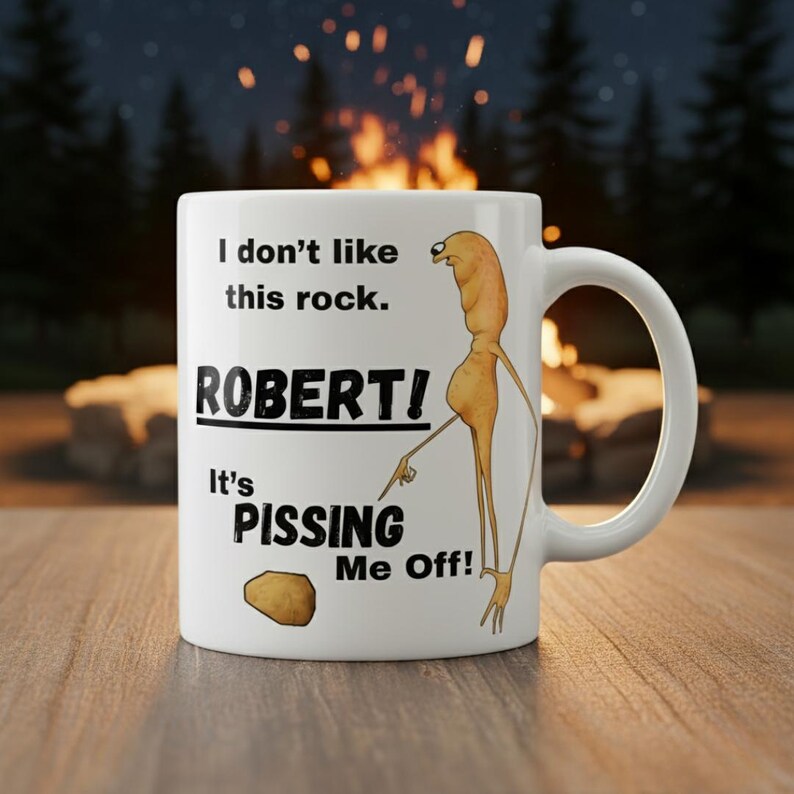 May include: White ceramic mug with a humorous design. The mug features the text "I don't like this rock. ROBERT! It's PISSING ME OFF!" along with a cartoon illustration of a figure pointing at a rock. The mug has a curved handle.