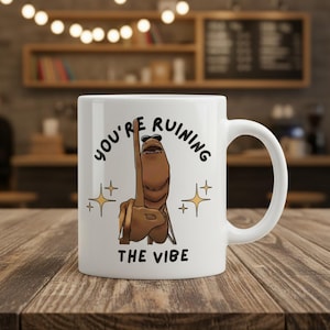 May include: White ceramic mug with a humorous design. The mug features the text "YOU'RE RUINING THE VIBE" with a cartoon illustration of a hand giving the middle finger. The design includes star accents.