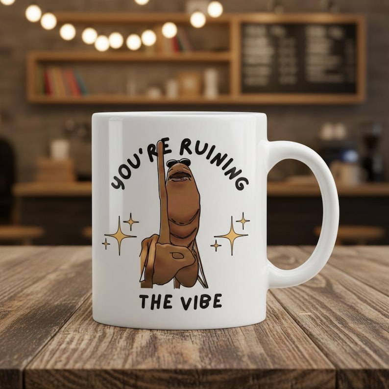 May include: White ceramic mug with a humorous design. The mug features the text "YOU'RE RUINING THE VIBE" with a cartoon illustration of a hand giving the middle finger. The design includes star accents.