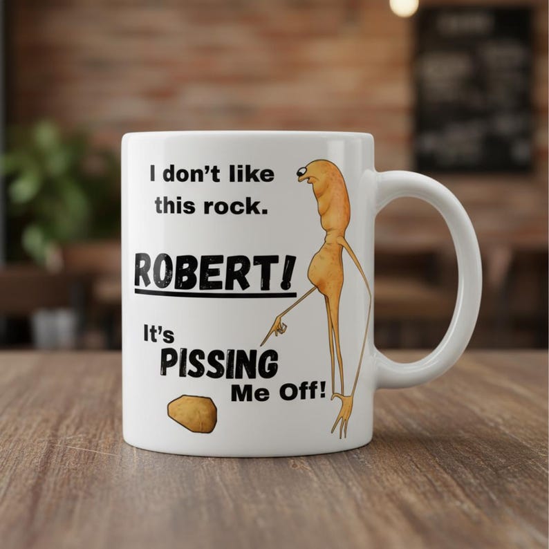 May include: White ceramic mug with a humorous design. The mug features the text "I don't like this rock. ROBERT! It's PISSING Me Off!" along with a cartoon illustration of a figure pointing at a rock. The mug has a curved handle.