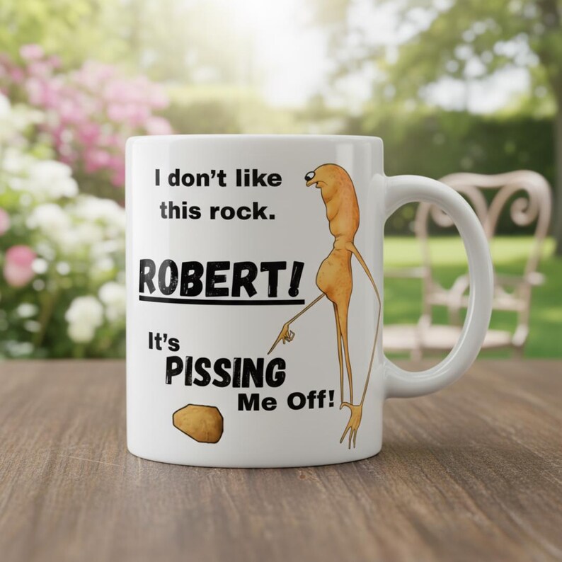 May include: White ceramic mug with a humorous design. The mug features the text "I don't like this rock. ROBERT! It's PISSING ME OFF!" along with a cartoon figure pointing at a rock. The mug has a curved handle.