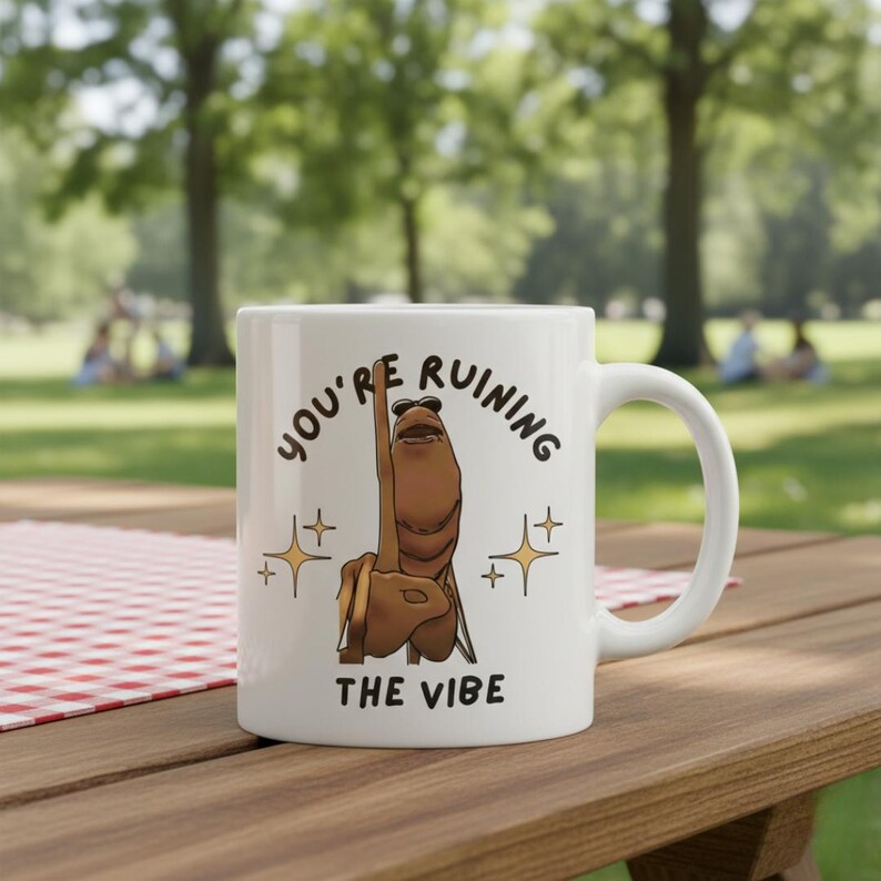 May include: White ceramic mug with a humorous design. The mug features a cartoon character giving the middle finger, with the text "YOU'RE RUINING THE VIBE" and star accents. The mug is on a wooden table.