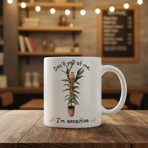 May include: White ceramic mug with a humorous design. The mug features a cartoon plant with the text "Don't yell at me... I'm sensitive." The plant is in a brown pot. The mug has a curved handle and is on a wooden table.