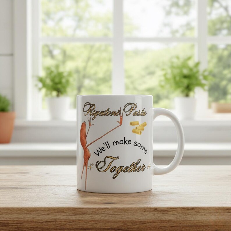 May include: White ceramic mug with the text "Rigatoni Pasta" and "We'll make some Together" in gold script. The mug features a whimsical illustration of a pasta figure. The mug is on a wooden surface.