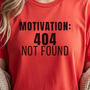 May include: A red t-shirt with the text "MOTIVATION: 404 NOT FOUND" in black. The short-sleeved shirt is made of a soft material. The person is wearing blue jeans and a beige cardigan.