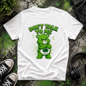 Pode incluir: Uma t-shirt branca de manga curta com o gráfico de um urso verde rabugento de braços cruzados. O urso tem uma mancha branca na barriga com um ícone de nuvem de chuva verde-escura. Acima do urso, o texto diz DON'T TALK TO ME em letras verdes.