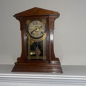 May include: A wooden antique clock with a dark brown finish. The clock face is white with black Roman numerals and gold accents. The clock has a pendulum and a decorative design.