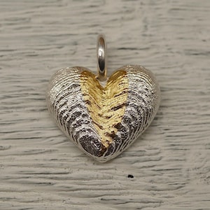 May include: A heart-shaped pendant with a textured silver exterior and a gold-colored center. The pendant has a small silver loop at the top for a chain. The design is detailed and the pendant is set against a light-colored wooden surface.