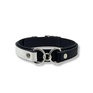 May include: A black and white leather collar with silver-tone metal rings. The collar is split into two sections, one white and one black, connected by two interlocking rings. The collar has a simple, elegant design.